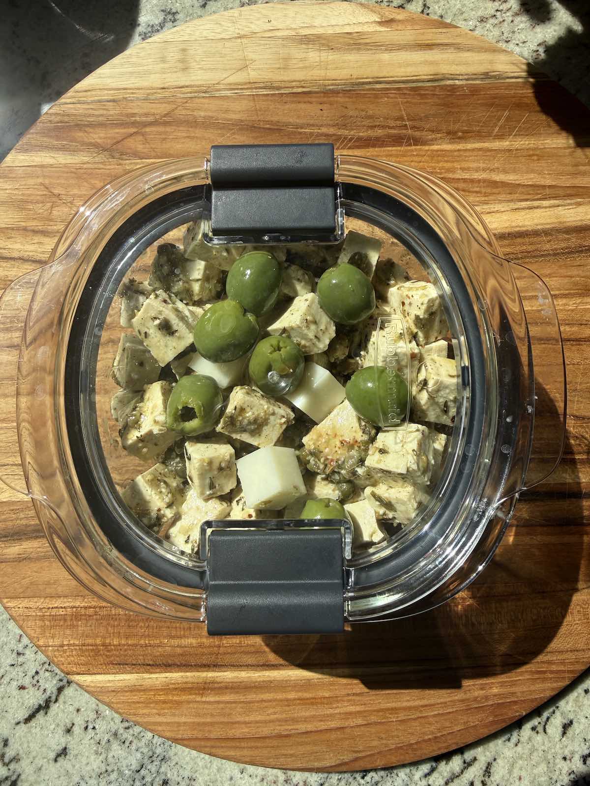 marinated tofu shown in covered dish with olives and cubes of cheese, ready to store in refrigerator.