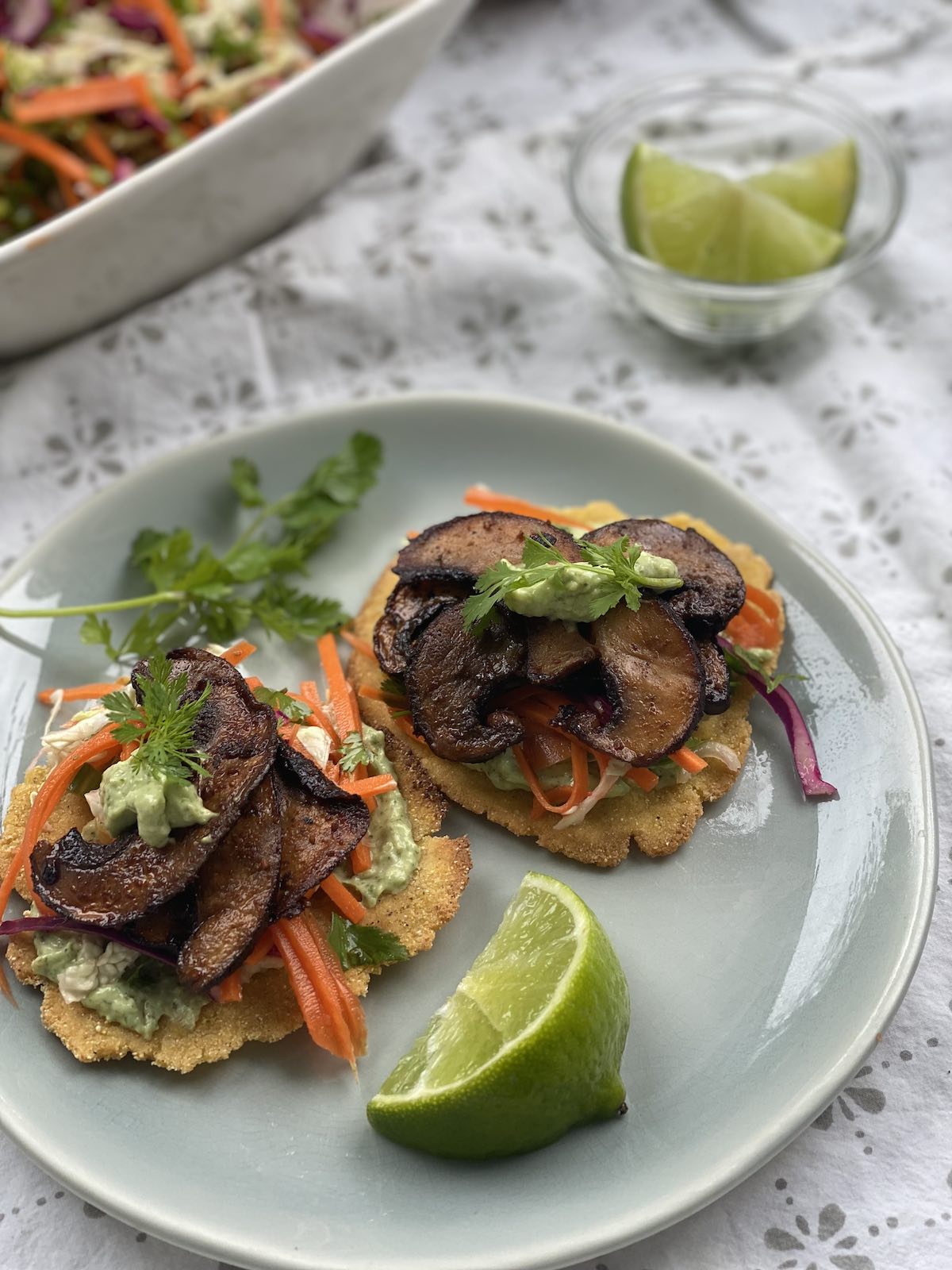 a serving of two mini tostadas on a plate with wedges of lime and bowl of extra slaw nearby.