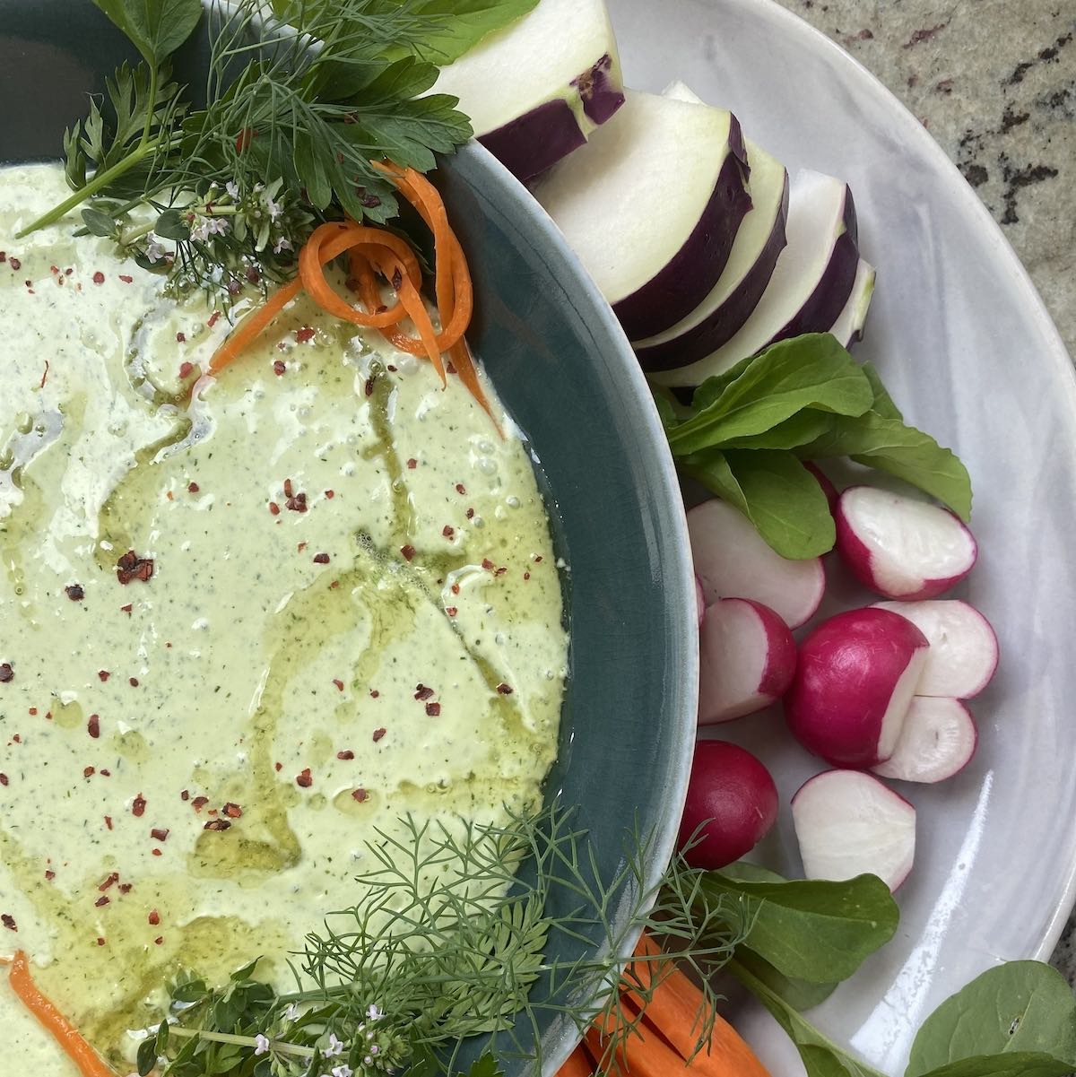 close up of whipped goat cheese dip garnished with fresh herbs, honey and Aleppo flakes and served with slices of carrot, radish and kohlrabi.