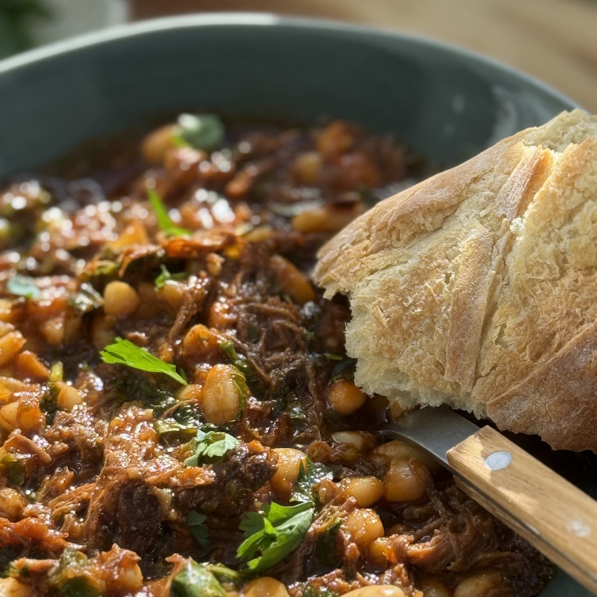 Serving of beef ragu with greens and beans, a piece of french bread on the side of the plate, and a spoon with a wooden handle.