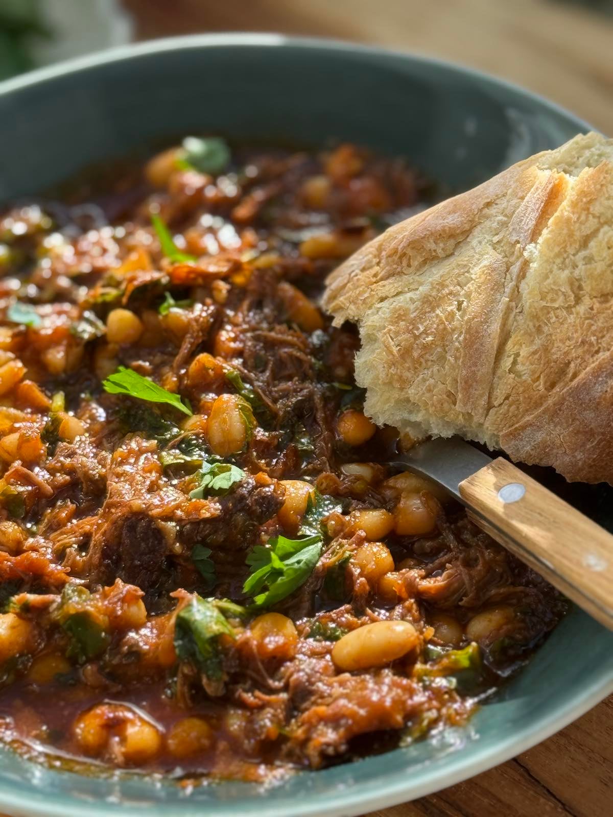 Serving of beef ragu with greens and beans, a piece of french bread on the side of the plate, and a spoon with a wooden handle.