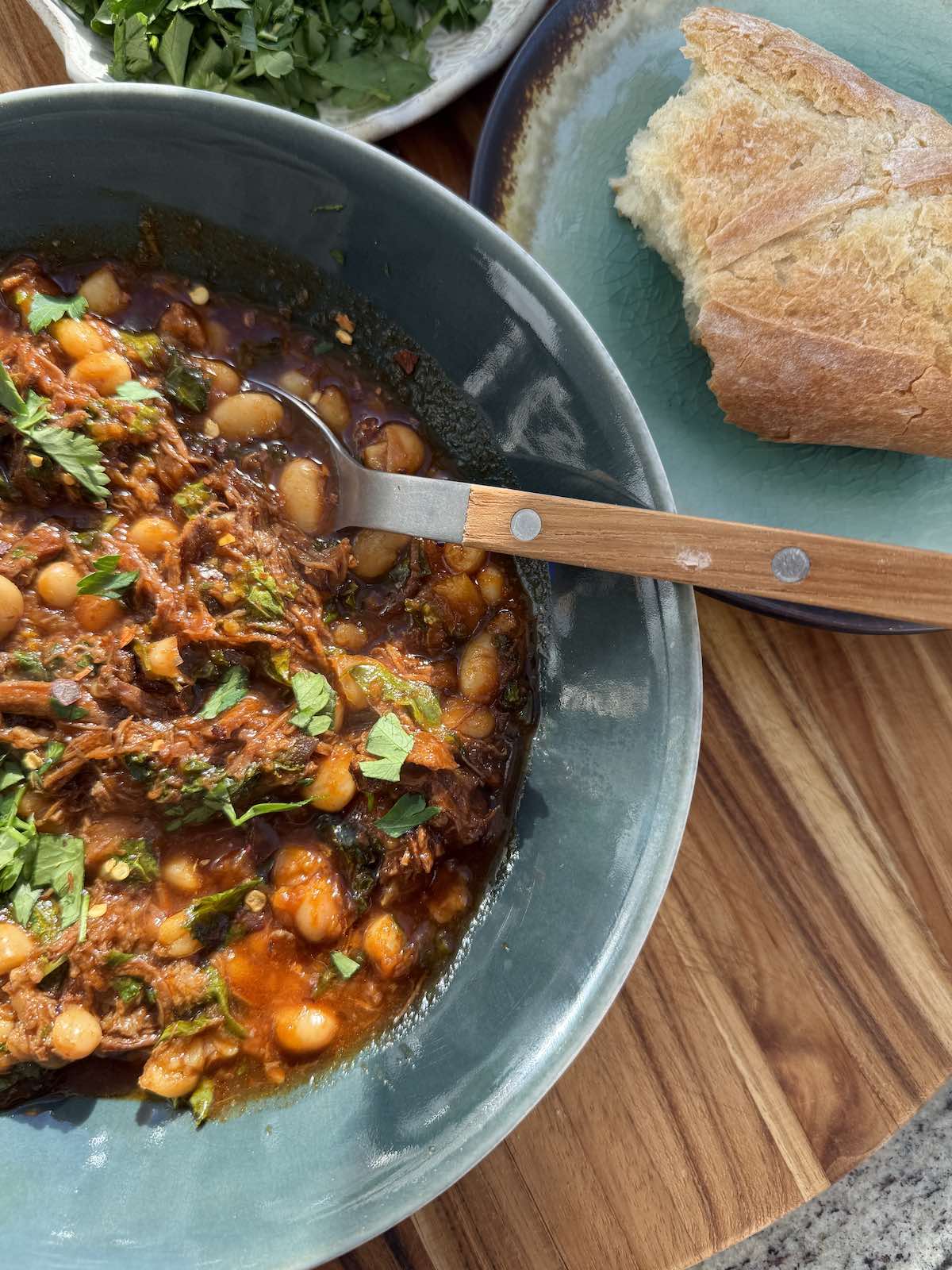 a close up of a serving of beef ragu with greens and beans, with a side of bread and parsley garnish.