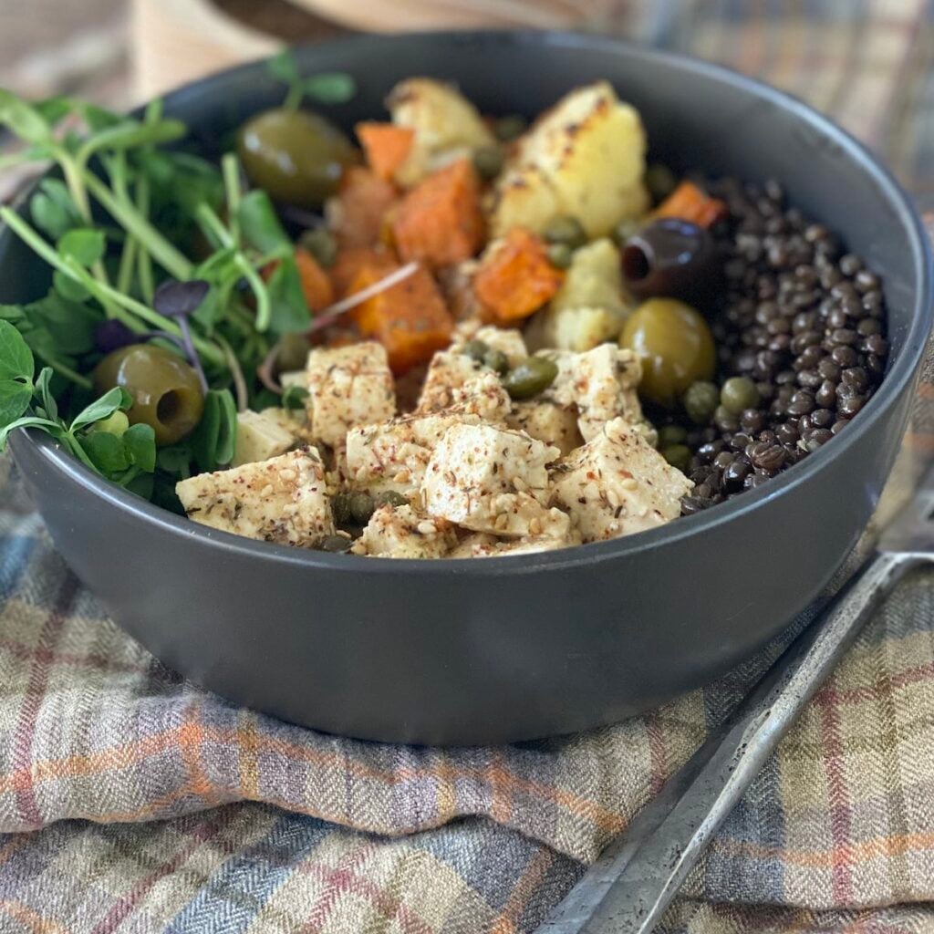 marinated tofu in a bowl with greens, olives, roasted veg and black lentils.