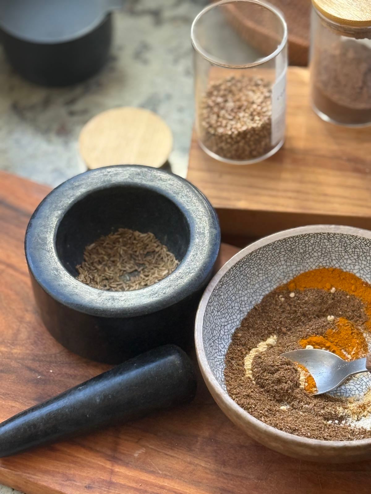 containers of whole and ground spices, a morter and pestle and a small bowl with several spices mixed together, all laid out on the counter.