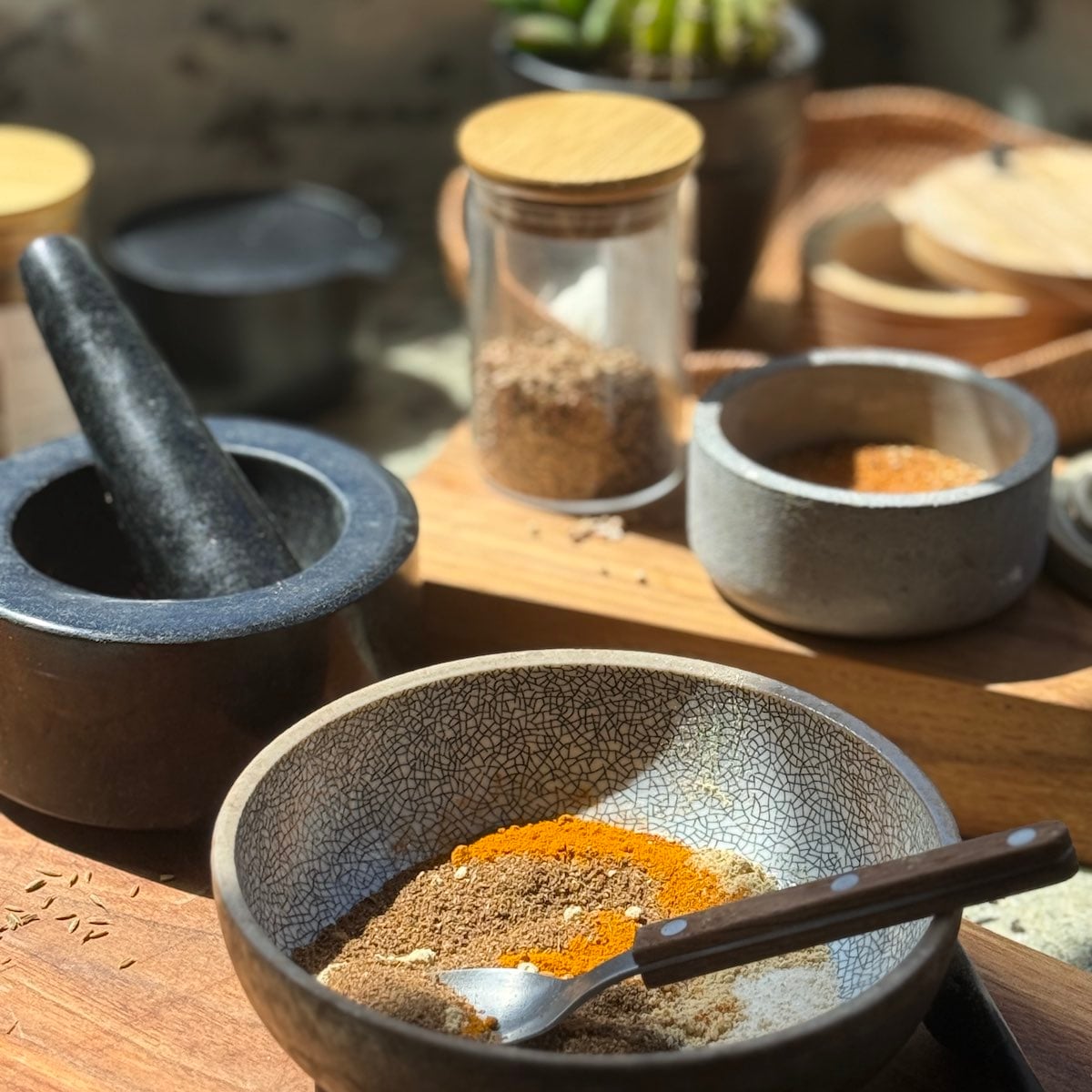 containers of whole and ground spices, a morter and pestle and a small bowl with several spices mixed together, all laid out on the counter.