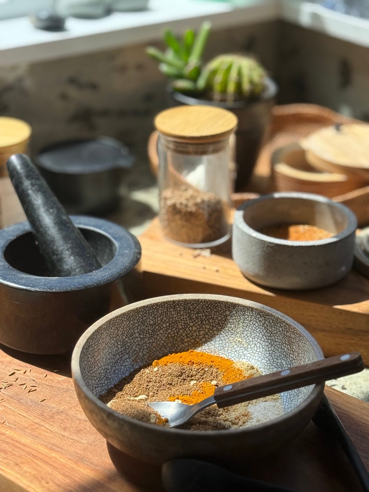 containers of whole and ground spices, a morter and pestle and a small bowl with several spices mixed together, all laid out on the counter.