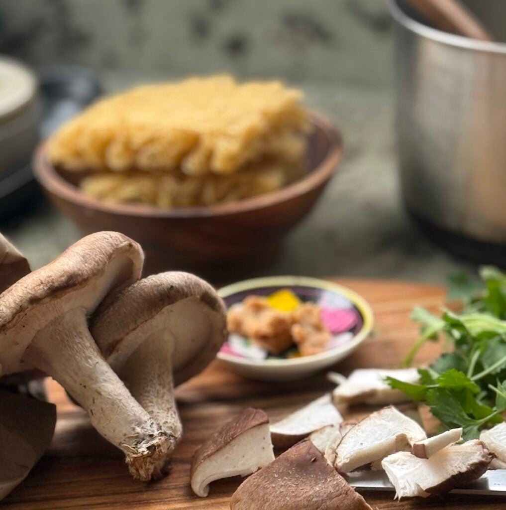 sliced shiitake mushrooms, two whole shiitakes, cilantro, miso and ramen set out on cutting board and counter, with soup pot and spoon nearby.