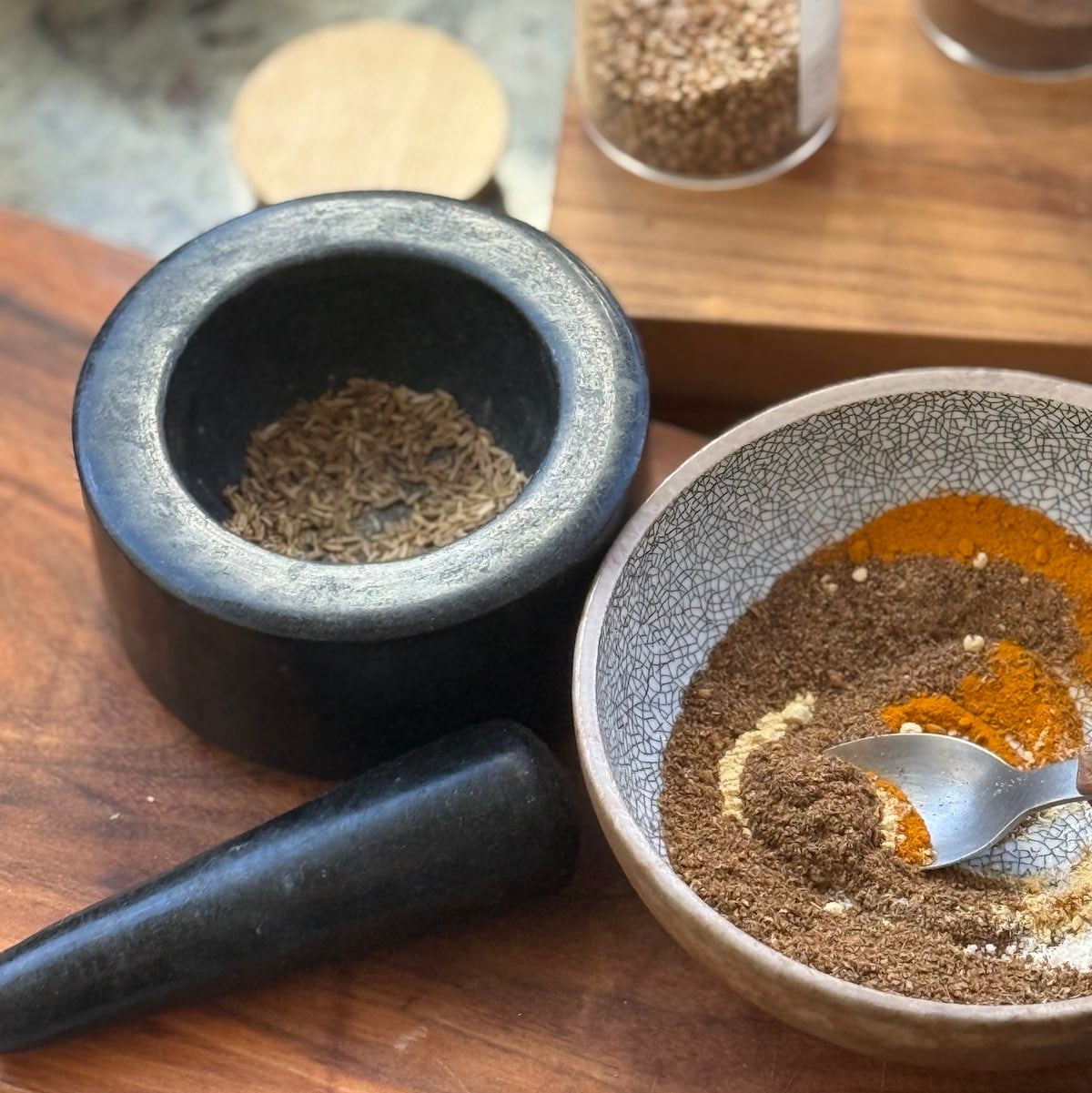 containers of whole and ground spices, a morter and pestle and a small bowl with several spices mixed together, all laid out on the counter.
