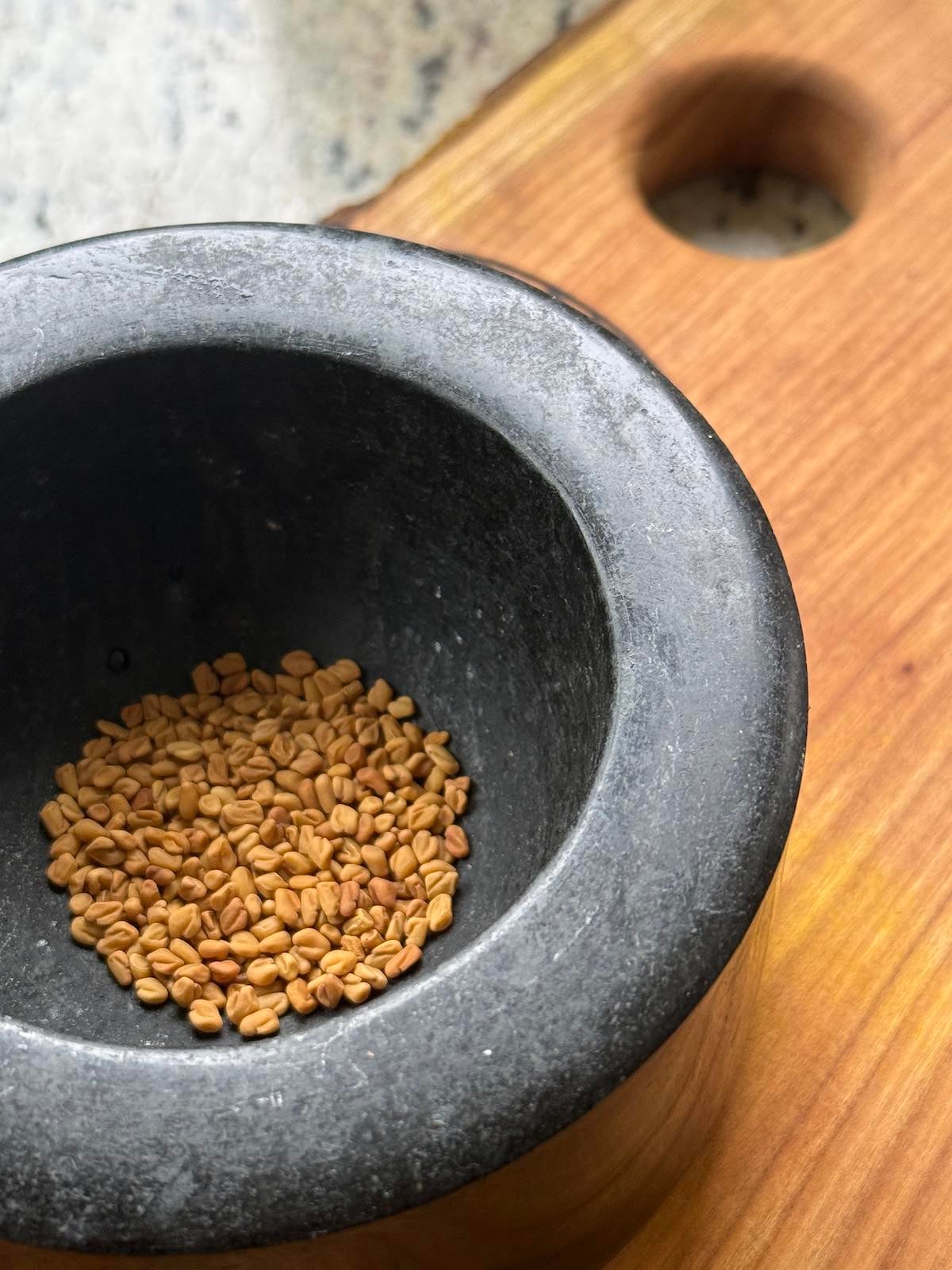 fenugreek seeds in a mortar, on a cutting board, ready to be ground with a pestle.