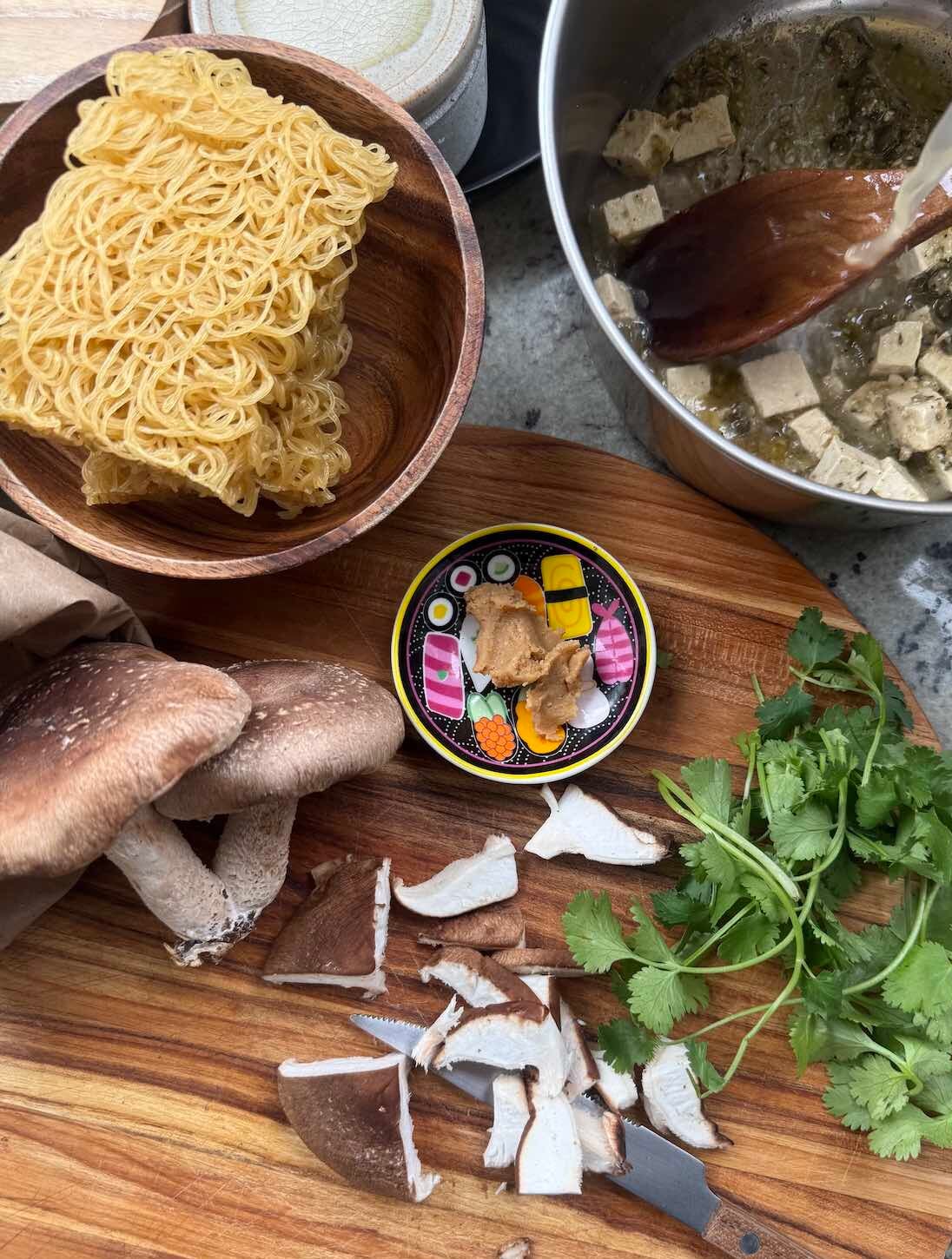 top down view of ramen soup ingredients, on cutting board, in process of being prepped, with soup pot and wooden spoon stirring in marinated tofu and broth.