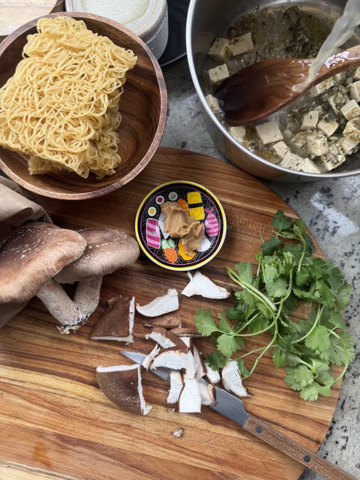 top down view of ramen soup ingredients, on cutting board, in process of being prepped, with soup pot and wooden spoon stirring in marinated tofu and broth.