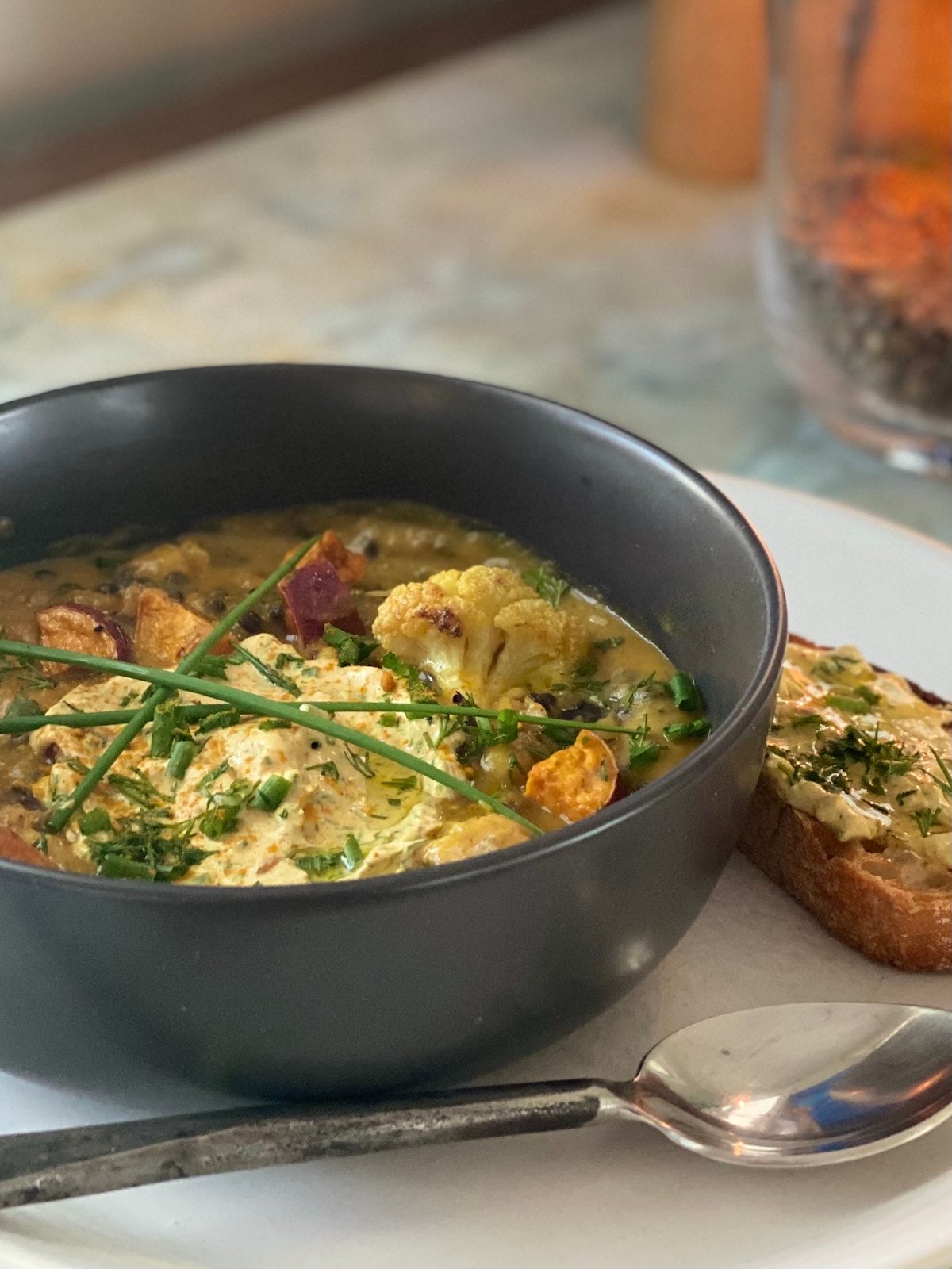 bowl of lentil, roasted vegetable soup, with spiced yogurt topping, chives, and a side of bread with yogurt topping, on a plate with a spoon.