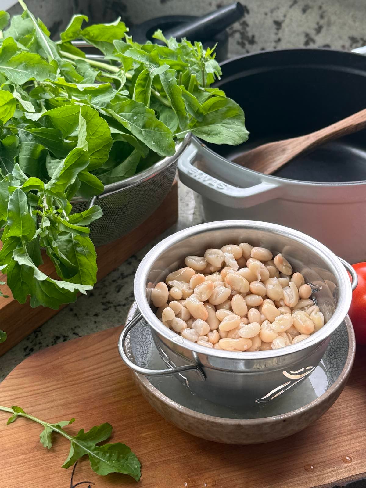 set up for making a meal, with wooden boards, a colander of beans and a colander of arugula, and a pot with a spoon in the background.