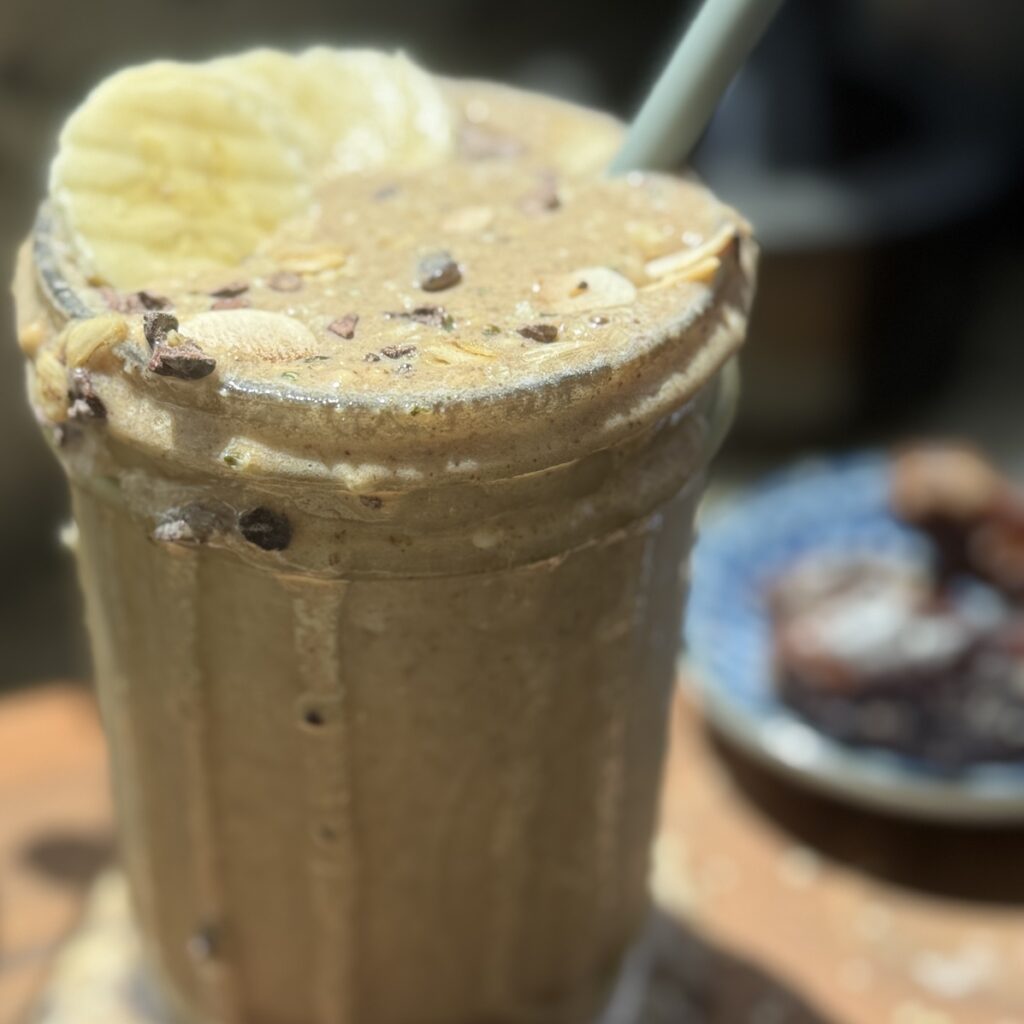 Blended espresso proteing smoothie with a straw, spilling over top of glass, and sliced banana on top with cacao nibs sprinkled over top.
