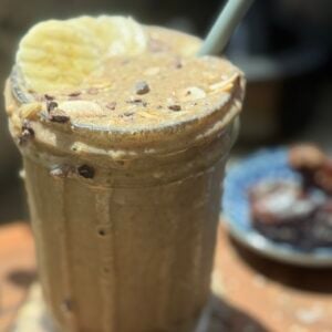 Blended espresso proteing smoothie with a straw, spilling over top of glass, and sliced banana on top with cacao nibs sprinkled over top.