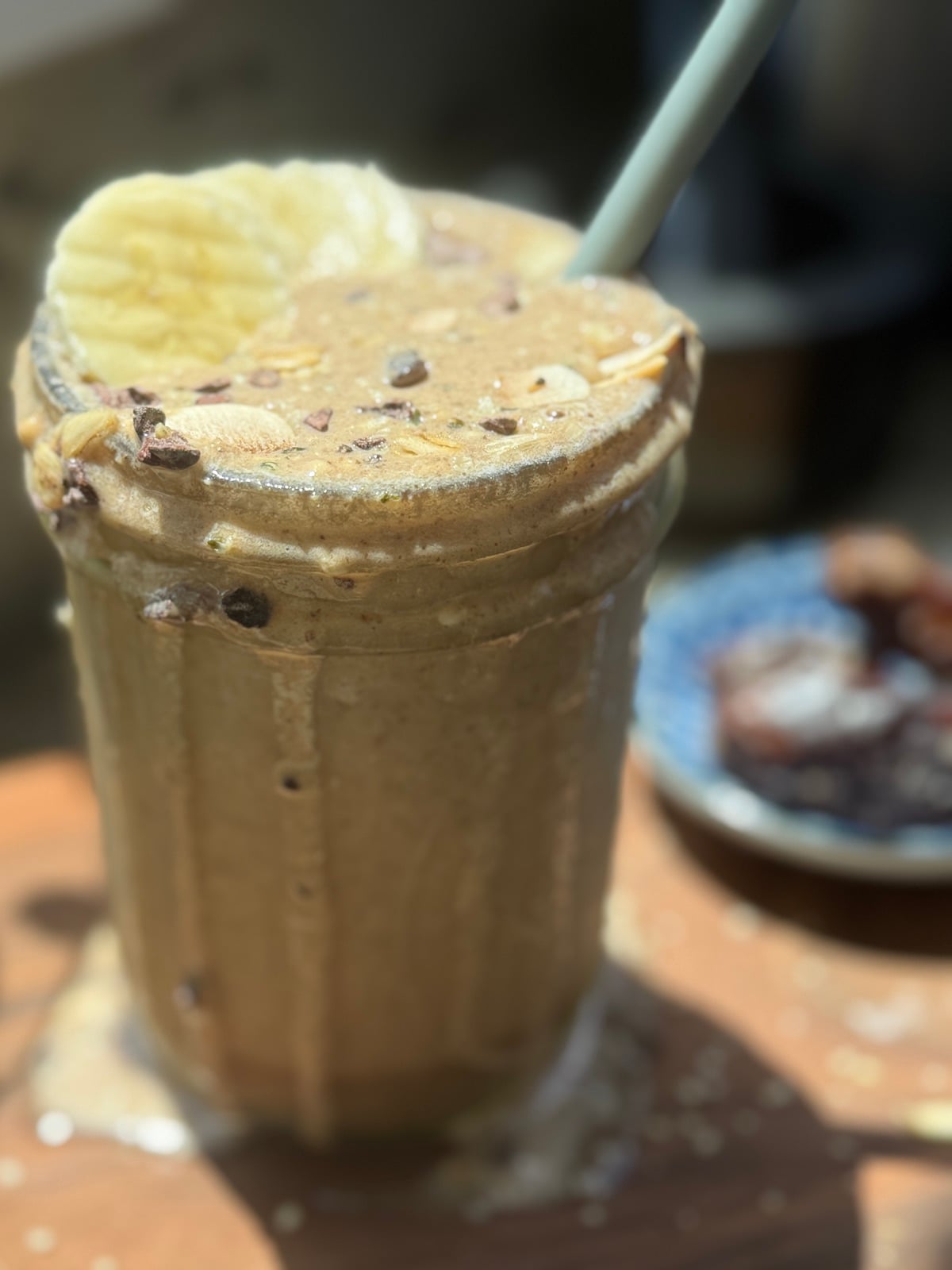 blended espresso proteing smoothie with a straw, spilling over top of glass, and sliced banana on top with cacao nibs sprinkled over top.